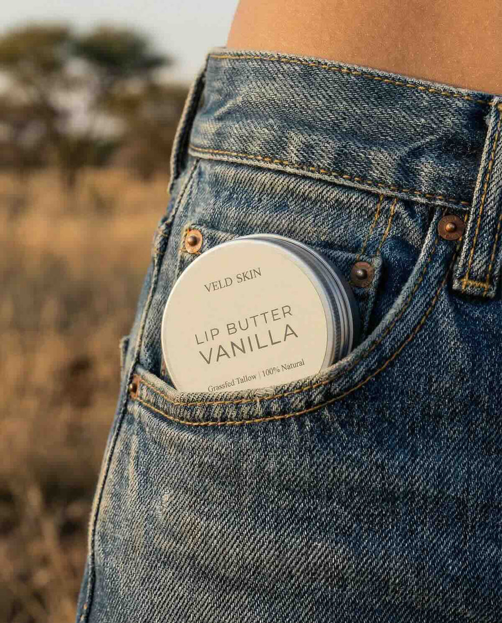 Lip butter container labeled 'Vanilla' in a jeans pocket with a blurred natural background