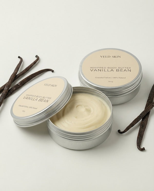 Vanilla bean whipped body butter in metal containers with vanilla beans on a light background