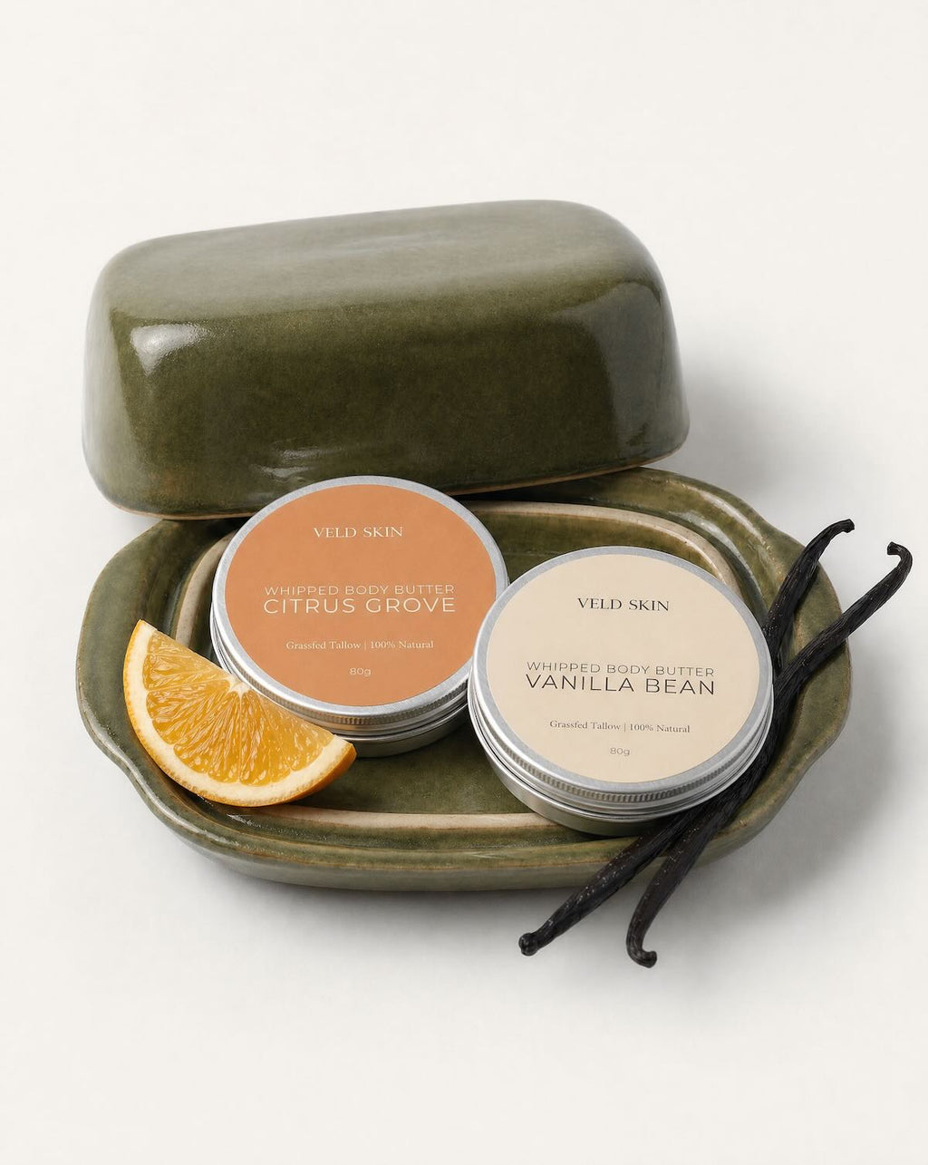 Two tins of Veld Skin body butter on a green tray with an orange slice and vanilla beans. 