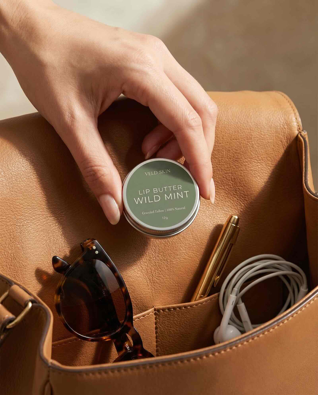 Hand placing a lip butter tin into a brown leather bag with sunglasses and earphones.