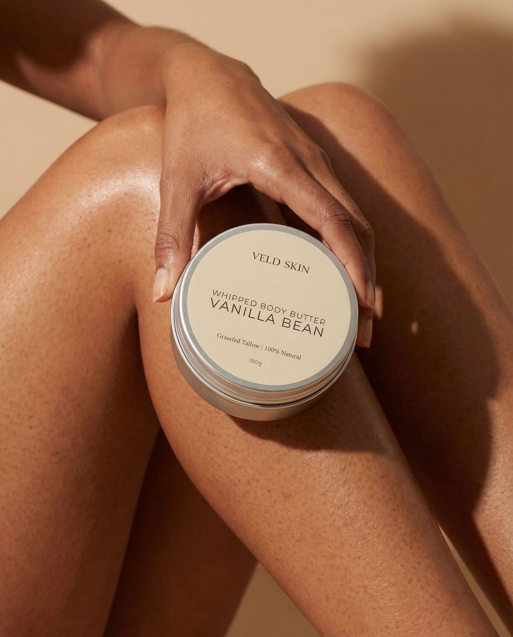 Hand holding a container of Veld Skin Vanilla Bean body butter against a beige background