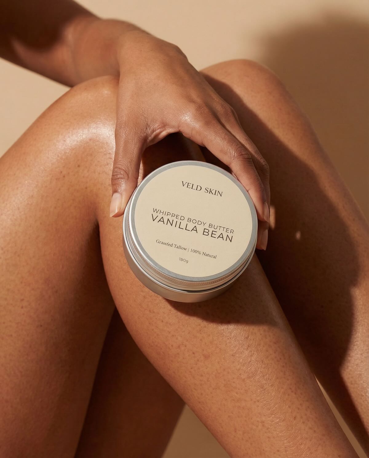 Hand holding a container of Veld Skin Vanilla Bean body butter against a beige background
