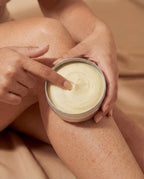 Person applying cream from a jar to their leg with a neutral background